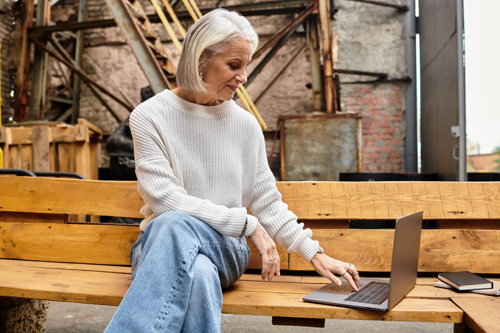 A mature woman working her weekend job on her laptop on a park bench.png